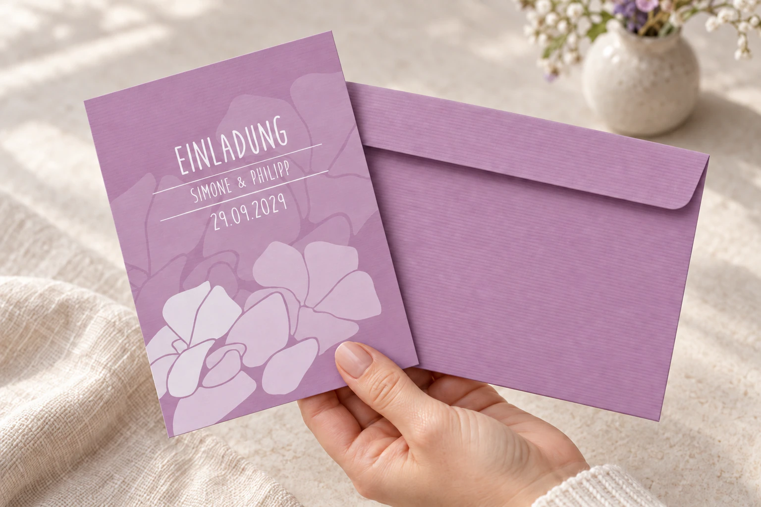Gerippte fliederfarbene Briefhülle und violette Hochzeitseinladung mit hellen Blüten in einer Hand